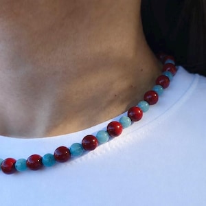Könnte beinhalten: Eine Perlenkette mit einem sich wiederholenden Muster aus runden, polierten dunkelroten Perlen und runden, hellblauen türkisfarbenen Perlen. Die Kette liegt um den Hals auf einem schlichten weißen T-Shirt und zeigt einen lebendigen Farbkontrast.
