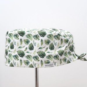 May include: A white scrub cap featuring a repeating pattern of various green monstera and fern leaves. The fabric head covering has a tie-back closure and a small white rectangular label on the side with the black text scrubdAU printed on it.