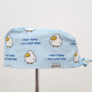 May include: A light blue scrub cap featuring a pattern of cartoon ducks and the text "I DON'T KNOW I JUST WORK HERE." The cap is tied at the back and has the scrubdAU logo.