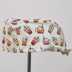 May include: A cream-coloured scrub cap featuring a repeating pattern of colourful bubble tea drinks with smiling faces. Each drink illustration includes a straw and boba pearls. A small white fabric tag with the text scrubdAU is sewn on the side.