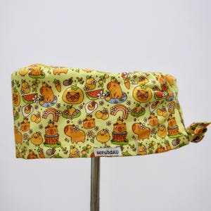 May include: A light green scrub cap featuring a repeating pattern of cartoon capybaras, rainbows, and food items like watermelon and cakes. The cap has a knotted tie in the back and the text "scrubdAU" is printed on the front.