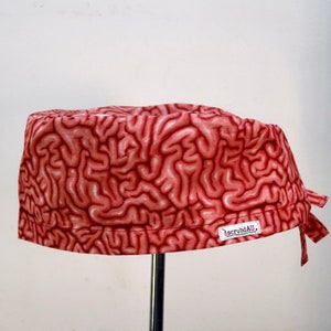 May include: A scrub cap featuring a repeating pattern of pink and red human brain tissue. The fabric has a detailed anatomical print of cerebral folds and sulci. A small white rectangular label with the black text scrubdAU is sewn on the side.