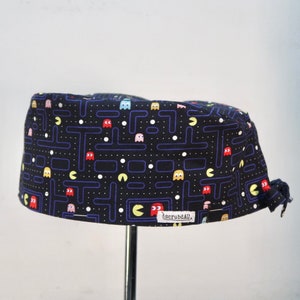 May include: A black scrub cap featuring a repeating pattern of yellow Pac-Man characters, colourful ghosts in red, pink, and light blue, and white dots against a dark blue maze grid. A small white fabric tag with the text scrubdAU is sewn on the side.