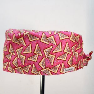 May include: A pink scrub cap featuring a repeating pattern of triangular toast slices topped with colourful sprinkles. The fabric cap has a tie-back closure and a small white rectangular label on the lower right side with the text scrubdAU.