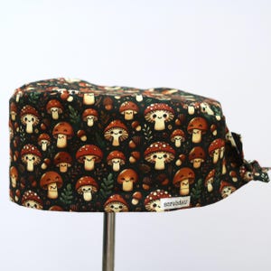 May include: A dark green scrub hat with a pattern of brown and orange mushrooms with white smiling faces. The hat has a small white label with the text "scrubAU".