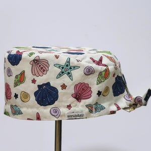 May include: A white scrub cap with a vibrant seashell design. The pattern includes pink, blue, and green seashells, starfish, and floral motifs. The text "scrubAU" is printed on the side.