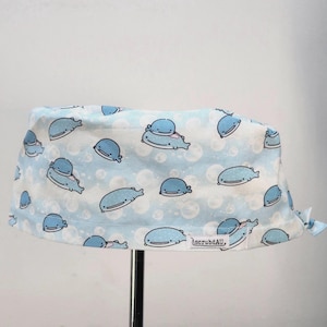 May include: A light blue scrub cap featuring a repeating pattern of cartoon whales and white bubbles. The fabric has a soft, pale blue background with various whale illustrations in different poses. A small white tag reads scrubdAU.