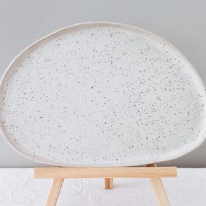 Könnte beinhalten: Weiße Keramik-Servierplatte mit gesprenkeltem Finish. Die Platte ist oval geformt und hat einen leicht erhöhten Rand. Sie wird auf einem Holzständer präsentiert.
