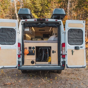 May include: A white van with the rear doors open, showing a custom built interior with a bed, storage shelves, and a bike rack. The van is parked in a woodland setting.
