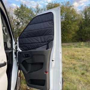 May include: A white van with a black window shade fitted in the side door. The shade is made of a thick, quilted material and has a black strap for easy opening and closing.