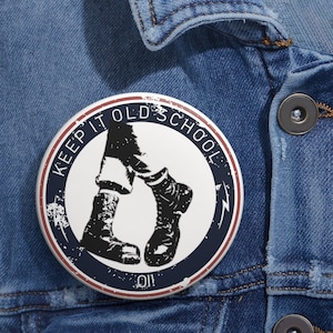 Könnte beinhalten: Ein weißer Knopf mit blauem und rotem Rand. Der Knopf hat einen abgenutzten Look und zeigt eine Schwarzweißillustration einer Person mit Stiefeln und dem Text "KEEP IT OLD SCHOOL OII!"