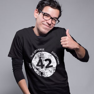 Puede incluir: Una camiseta negra de manga larga con un gráfico circular blanco que presenta el texto "Life, the Universe, and Everything" y el número "42" en el centro. La persona que lleva la camiseta sonríe y levanta el pulgar.