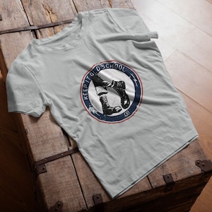 Könnte beinhalten: Hellgraues T-Shirt mit einem kreisförmigen Grafikdesign mit schwarz-weißen Stiefeln und dem Text "KEEP IT OLD SCHOOL" in einem blau-roten Design. Das T-Shirt wird auf einer Holzoberfläche präsentiert.