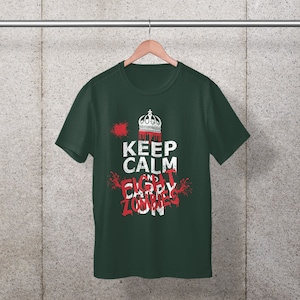 以下が含まれることがあります： 「Keep Calm and Fight Zombies」と白で書かれたグラフィックプリントが施された濃い緑色のTシャツ。グラフィックには王冠と赤いしぶきも含まれています。