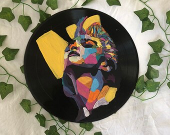 May include: A black vinyl record featuring a colorful abstract painting of a face. The artwork uses shades of yellow, purple, blue, and pink. Green ivy vines are arranged around the record on a white background.