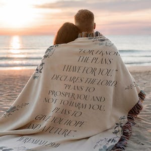 May include: A beige woven blanket with floral patterns and fringed edges, covering two people on a beach. The blanket has a printed quote from Jeremiah 29:11 in black text.