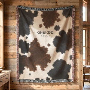 May include: A woven throw blanket featuring a brown and cream cowhide pattern with fringed edges. The center displays the text G&H above the date 4.9.2026 in a dark serif font. The blanket is rectangular with a textured surface and visible weave.