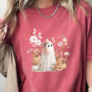 May include: Crimson-colored t-shirt featuring a watercolor illustration of a ghost with a bow, surrounded by pumpkins and flowers. The ghost has a white sheet with a lace trim. The shirt is short-sleeved and has a relaxed fit.