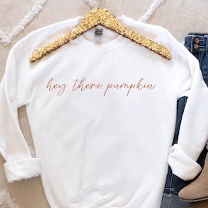May include: White sweatshirt with the text "hey there pumpkin" in orange lettering. The sweatshirt is laying on a white and beige rug with a pair of brown ankle boots and blue jeans.