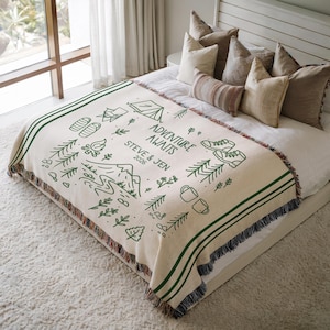 May include: Cream-colored throw blanket with green stripes and fringe. The blanket features a green line drawing of camping and hiking elements, with the text "ADVENTURE AWAITS" and names and a year.