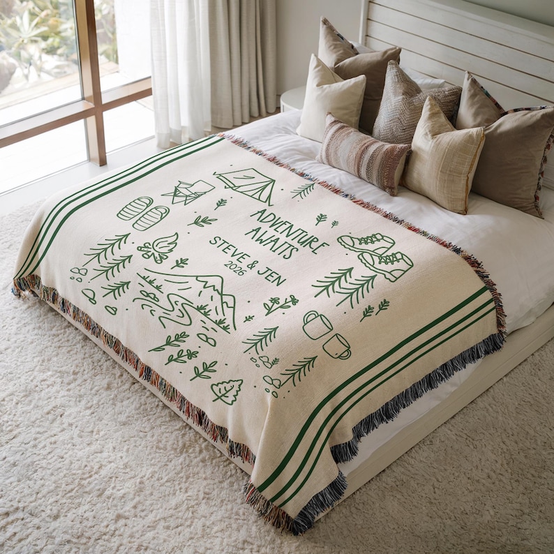 May include: Cream-colored throw blanket with green stripes and fringe. The blanket features a green line drawing of camping and hiking elements, with the text "ADVENTURE AWAITS" and names and a year.