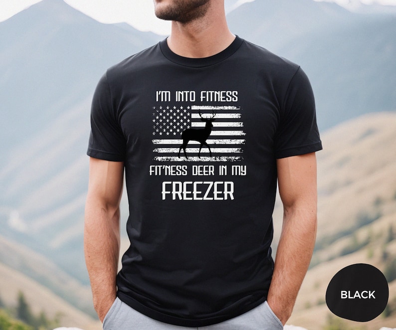 Puede incluir: Camiseta negra con un dise&ntilde;o de bandera estadounidense desgastada en blanco que presenta una silueta de ciervo. El texto "I'm into fitness fitness deer in my freezer" est&aacute; impreso debajo de la bandera.