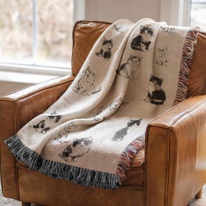 May include: A cream-colored woven throw blanket featuring a repeating pattern of black and white kitten illustrations. The blanket has a textured fabric finish and is finished with decorative fringe along the bottom edge.