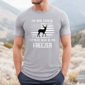 Puede incluir: Una camiseta gris con el texto "I'm into fitness fitness deer in my freezer" y una imagen de un ciervo frente a una bandera estadounidense.
