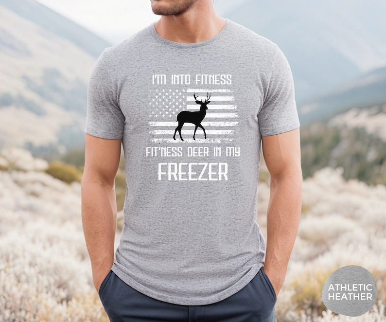 Puede incluir: Una camiseta gris con el texto "I'm into fitness fitness deer in my freezer" y una imagen de un ciervo frente a una bandera estadounidense.