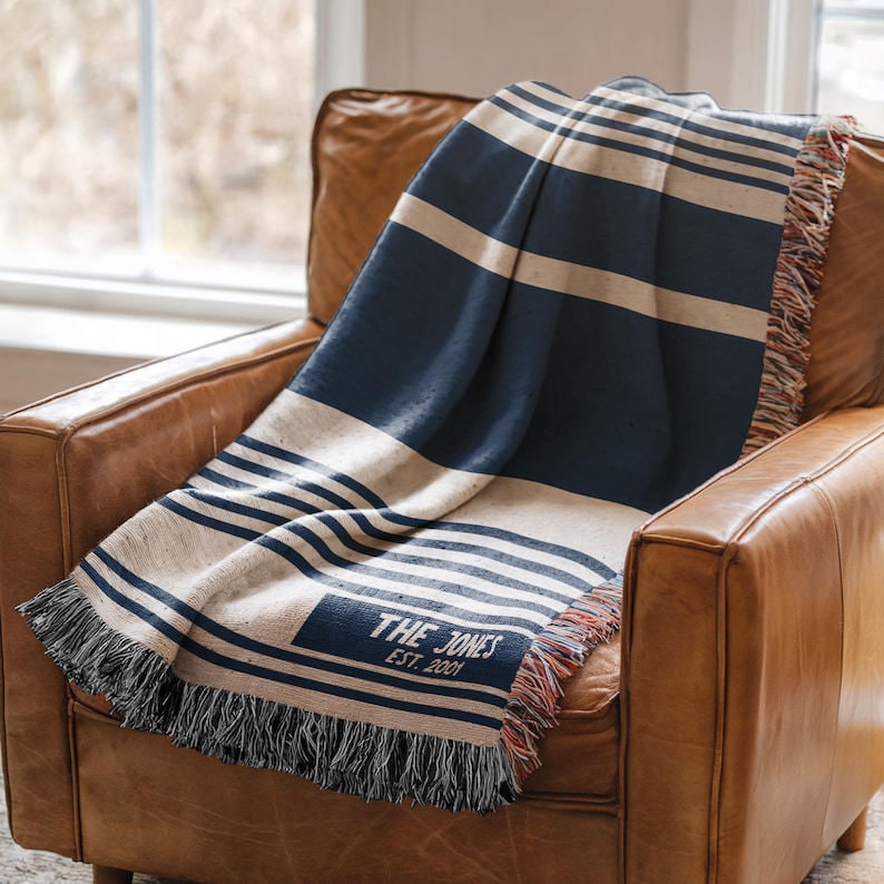 May include: A navy blue and cream striped throw blanket draped over a brown leather armchair. The blanket has a fringed edge and the text "THE JONES EST. 2001" is visible on the corner.