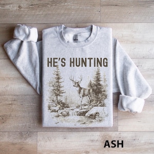 May include: Ash-colored sweatshirt with the text "HE'S HUNTING" in brown. The graphic depicts a deer standing near a stream with trees in the background. The word "ASH" is printed at the bottom.