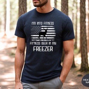Puede incluir: Una camiseta azul marino con un dise&ntilde;o de bandera estadounidense desgastado que presenta una silueta de ciervo. El texto "I'm into fitness fitness deer in my freezer" est&aacute; impreso en blanco en la camiseta.