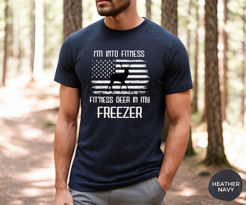 Puede incluir: Una camiseta azul marino con un dise&ntilde;o de bandera estadounidense desgastado que presenta una silueta de ciervo. El texto "I'm into fitness fitness deer in my freezer" est&aacute; impreso en blanco en la camiseta.