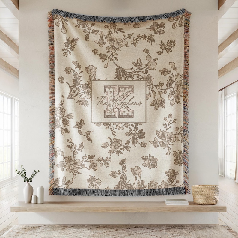 Op de afbeelding: Cr&egrave;mekleurige bloemen plaid met een decoratieve rand en franjes. De deken heeft een herhalend patroon van bloemen en bladeren in bruine tinten. De tekst "The Kaplans" wordt weergegeven in een rechthoekig kader.