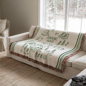 May include: Cream-colored woven throw blanket with a green illustrated camping design. The blanket features a tent, hiking boots, trees, and the words "ADVENTURE AWAITS" and "STEVE & JEN". Green stripes and fringe trim the edges.
