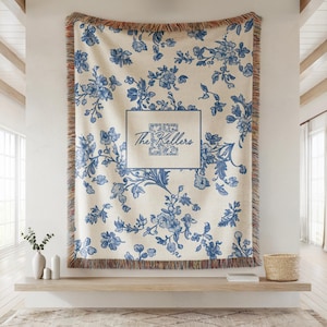 May include: A cream-colored throw blanket with a blue floral pattern and fringed edges. The center features a framed design with the text "The Kellers". The blanket is displayed on a wall, adding a decorative touch to the room.