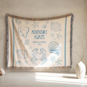 May include: Beige woven throw blanket with blue illustrations of seashells, starfish, and a crab. The text "ADVENTURE AWAITS" is centered, with names and a year below. The blanket has a fringed edge and blue stripes.