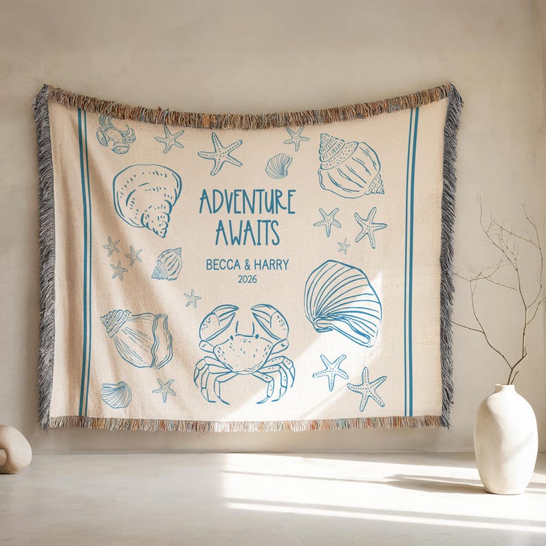 May include: Beige woven throw blanket with blue illustrations of seashells, starfish, and a crab. The text "ADVENTURE AWAITS" is centered, with names and a year below. The blanket has a fringed edge and blue stripes.