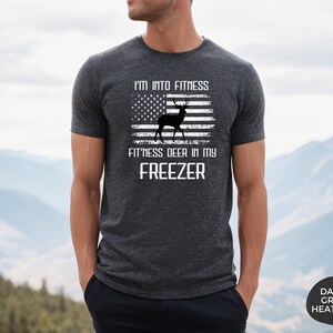Puede incluir: Una camiseta de color gris oscuro jaspeado con un dise&ntilde;o de bandera estadounidense desgastado que presenta una silueta de ciervo. El texto dice "I'm into fitness fitness deer in my freezer".