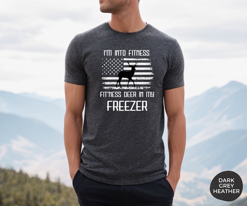 Puede incluir: Una camiseta de color gris oscuro jaspeado con un dise&ntilde;o de bandera estadounidense desgastado que presenta una silueta de ciervo. El texto dice "I'm into fitness fitness deer in my freezer".