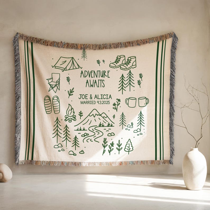 May include: Cream-colored woven throw blanket with a green camping-themed design. Features illustrations of a tent, hiking boots, trees, and the text "ADVENTURE AWAITS" with a personalized name and wedding date. The blanket has green stripes and fringe.
