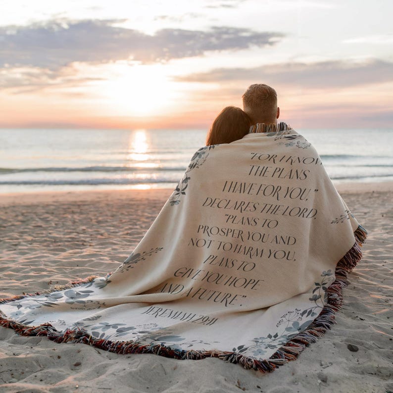 May include: A beige woven blanket with floral accents and fringed edges, draped over two people on a beach. The blanket features a printed quote from Jeremiah 29:11 in black text.