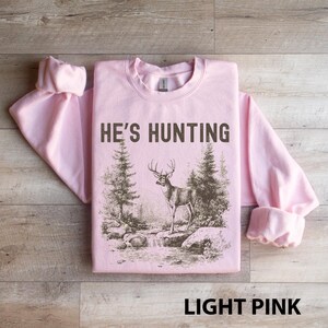 May include: Light pink sweatshirt with the text "HE'S HUNTING" in dark brown. The graphic features a deer standing near a stream with trees. The words "LIGHT PINK" are at the bottom.