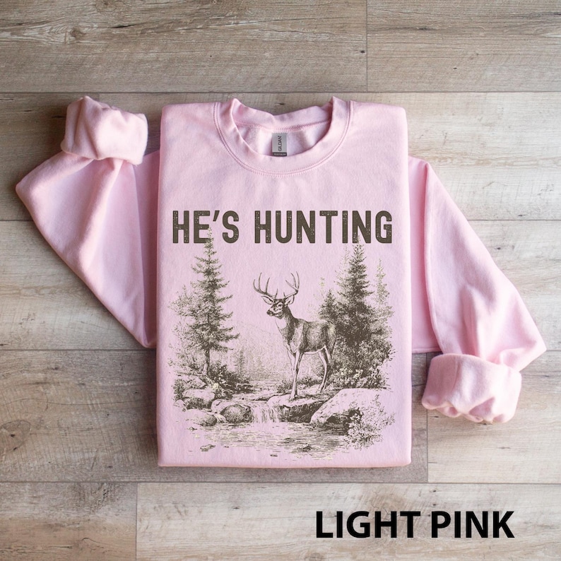 May include: Light pink sweatshirt with the text "HE'S HUNTING" in dark brown. The graphic features a deer standing near a stream with trees. The words "LIGHT PINK" are at the bottom.