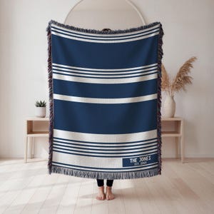 May include: A navy blue and white striped throw blanket with a fringed edge. The blanket has horizontal white stripes and a rectangular label with the text "THE JONES EST. 2001". The blanket is approximately 60 inches wide and 80 inches long.