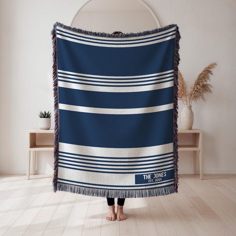 May include: A navy blue and white striped throw blanket with a fringed edge. The blanket has horizontal white stripes and a rectangular label with the text "THE JONES EST. 2001". The blanket is approximately 60 inches wide and 80 inches long.
