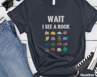 May include: A heather navy short-sleeve t-shirt featuring the white text WAIT I SEE A ROCK above a grid of sixteen colorful illustrations of various rocks, minerals, and crystals in shades of blue, green, yellow, orange, and grey.