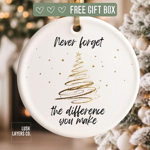 May include: A white ceramic Christmas ornament with gold text and a gold ribbon. The ornament features a gold Christmas tree design and the words "Never forget the difference you make."