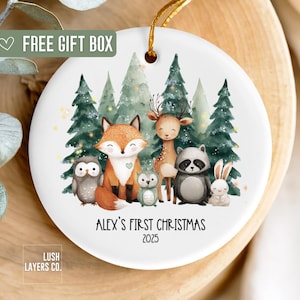 May include: A white ceramic Christmas ornament featuring a woodland scene with a fox, deer, owl, raccoon, and rabbit. The text "Alex's First Christmas 2025" is printed below the animals. Includes a gold string for hanging.