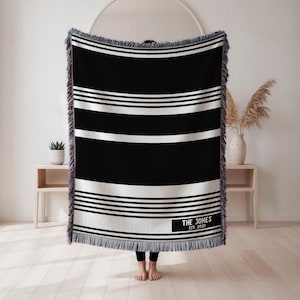 May include: A black and white striped throw blanket with a rectangular label that reads "THE JONES EST. 2001". The blanket has a fringed edge and is displayed in front of a white wall.